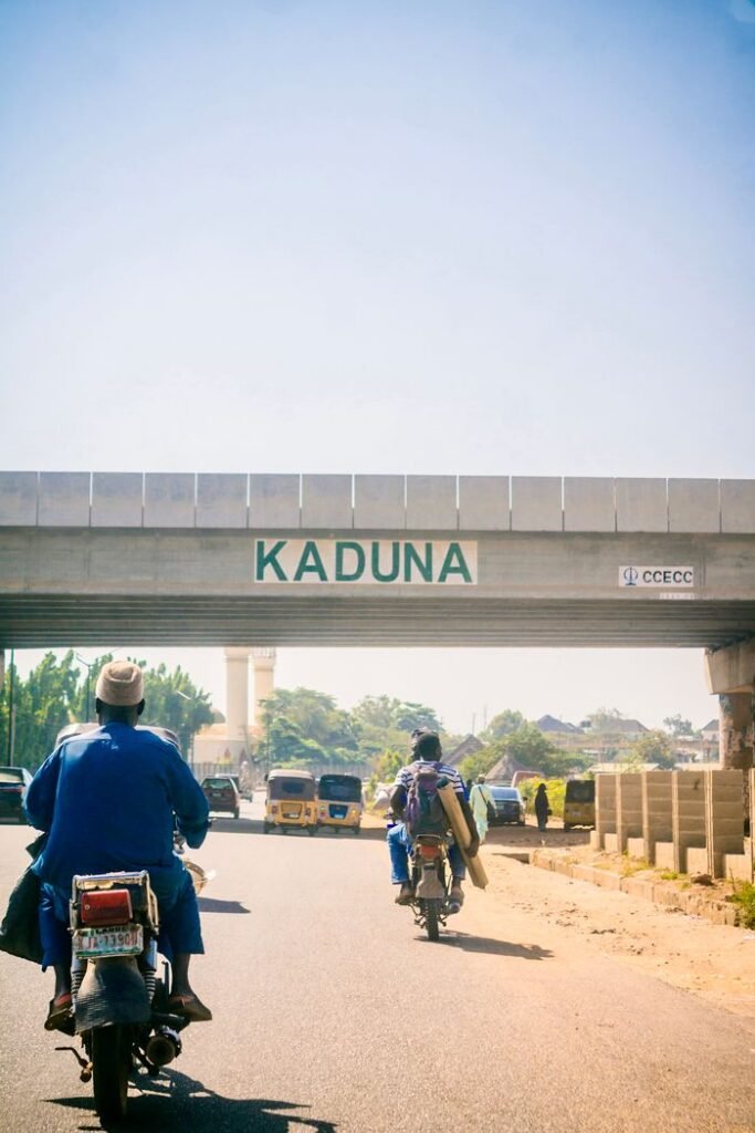 Motorcyclists travel under bridge marked 'Kaduna' in sunny Nigeria.
